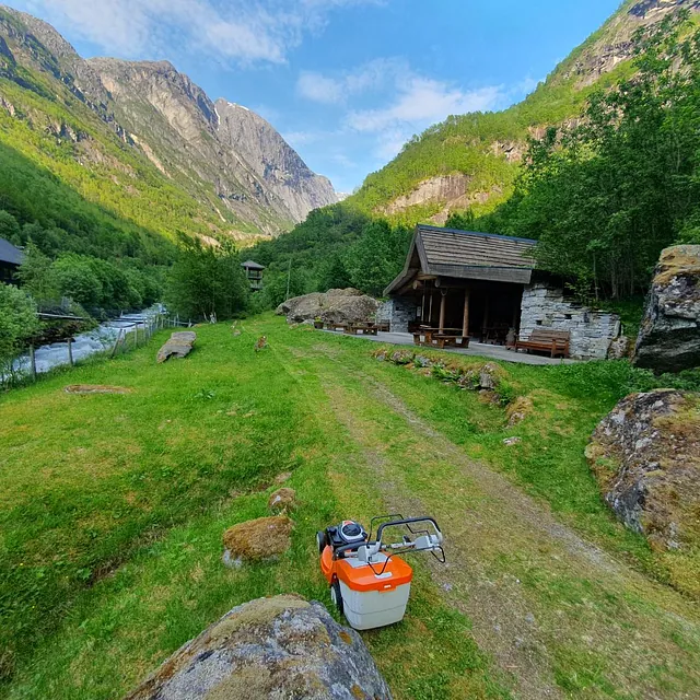 Gressplen og hytte omringet av fjell, med liten elv i bakgrunnen.