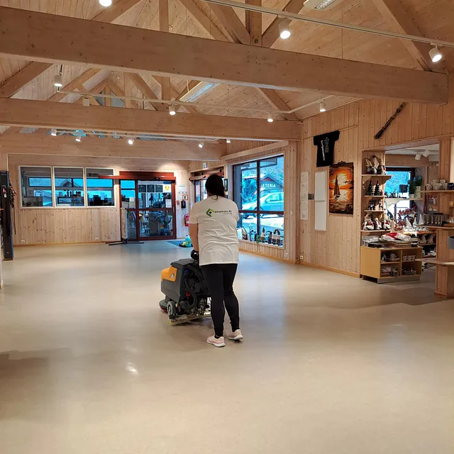 Person vasker gulvet med gulvvaskemaskin i romslig butikk med trebjelker.