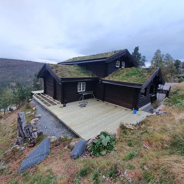 Hytte med torvtak og stor terrasse, omgitt av fjellandskap og høstfarger.