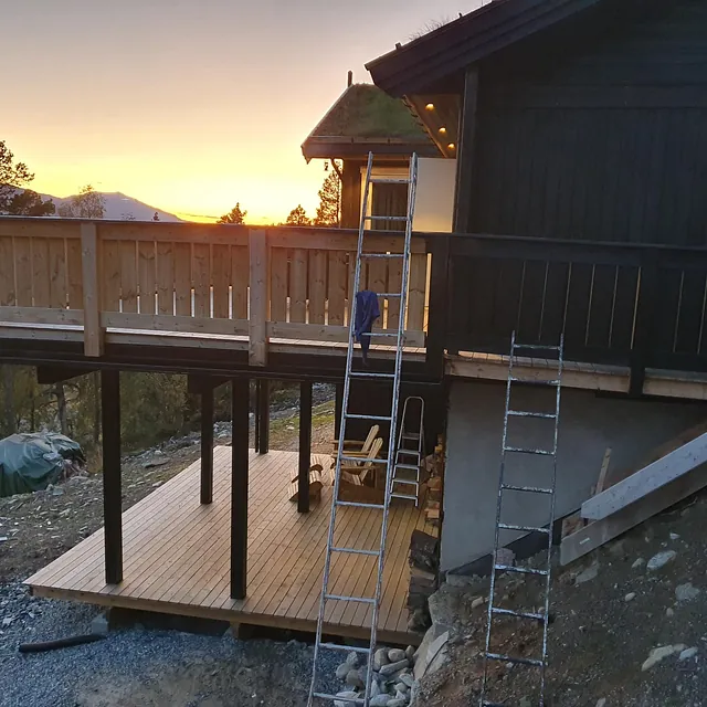 Terrasse i tre med solnedgang i bakgrunnen, stillas og stige plassert ved bygget.